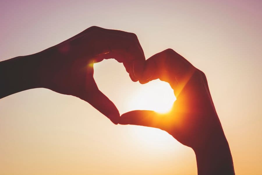 Two hands raised against a sunset sky, forming the shape of a heart.
